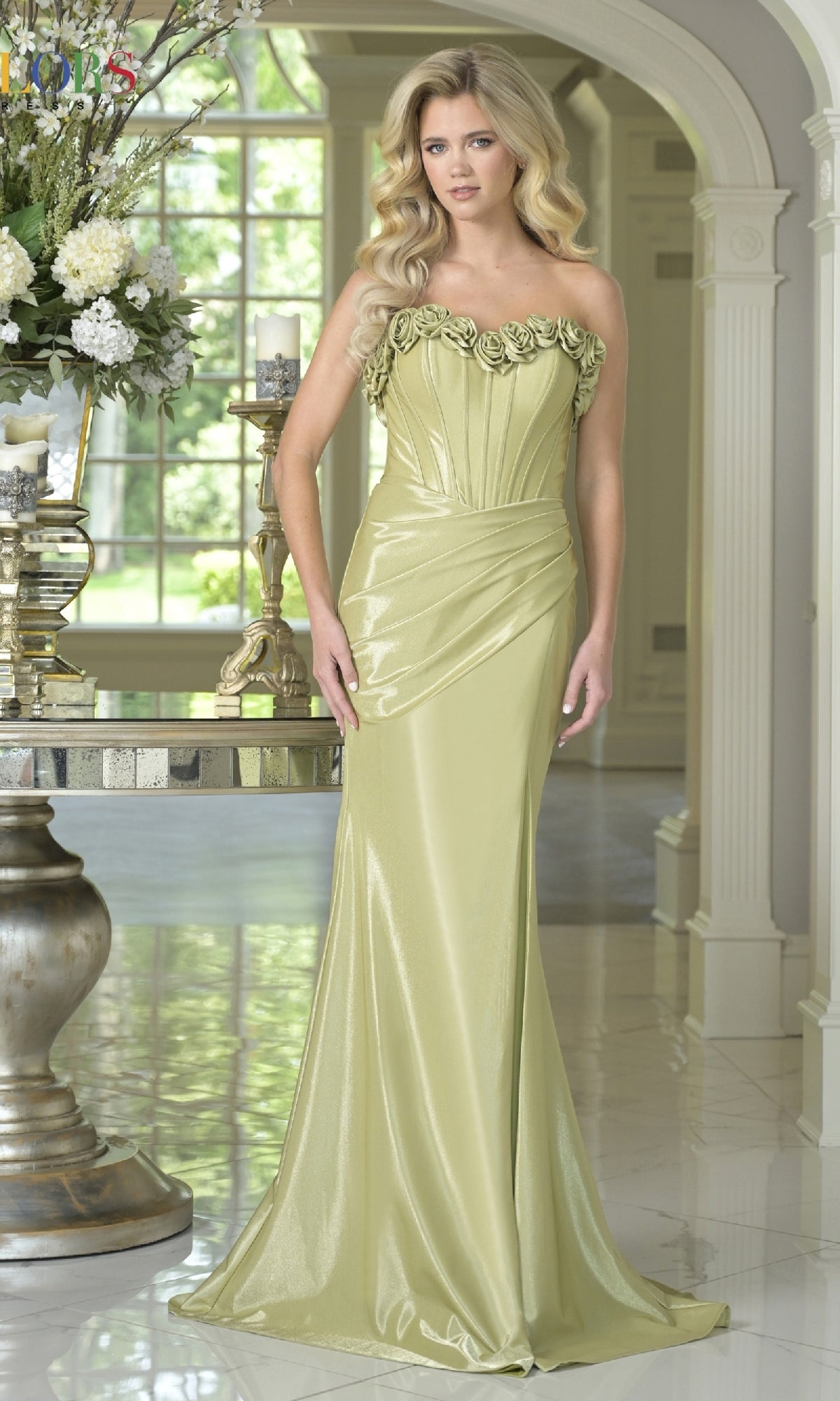 Woman wearing the Strapless Long Rosette Prom Gown (Colors Dress 3895) in sage, featuring a sweetheart neckline and floral bodice, stands in an elegant, bright room with ornate decor and large windows.